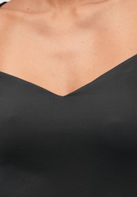 Moteriška viršutinė liemens dalis, apsirengusi aptempta juoda palaidine be petnešų, rodančia kaklo iškirpimą ir viršutinę krūtinės sritį.