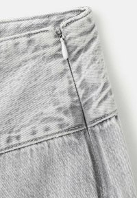 Primer plano de tela de mezclilla gris claro que muestra una cremallera metálica parcialmente cerrada con una pestaña blanca de tirador cosida en la costura.