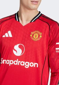 Das rote Manchester United Trikot hat einen V-Ausschnitt, schwarze Akzente und Logos: Adidas auf der Schulter, Snapdragon auf der Vorderseite und das Vereinswappen.