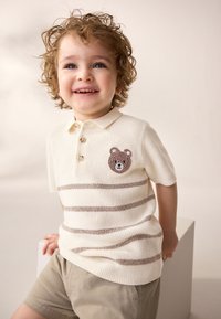 Koszulka polo z krótkim rękawem w kolorze kremowym z brązowymi paskami, z haftem twarzy misia na piersi. Do tego beżowe szorty.