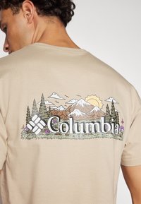 Tricou bej cu un imprimeu colorat de peisaj montan, cu copaci, flori și logo-ul "Columbia" afisat proeminent.