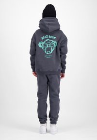 Persoon draagt een donkergrijze hoodie en joggingbroek, zwarte muts, witte sneakers, staat met gezicht naar achteren gericht met een grafisch ontwerp op de rug van de hoodie.