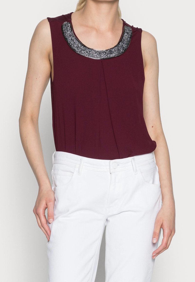Femme portant un haut sans manches rouge foncé avec un col orné de perles argentées et un pantalon blanc, debout devant un fond uni.