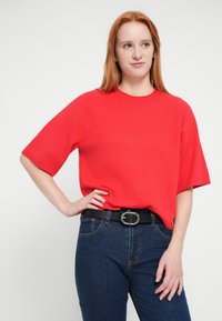 Top rojo de punto de manga corta con cuello redondo, combinado con vaqueros de mezclilla azul oscuro de talle alto y un cinturón negro con una hebilla plateada.