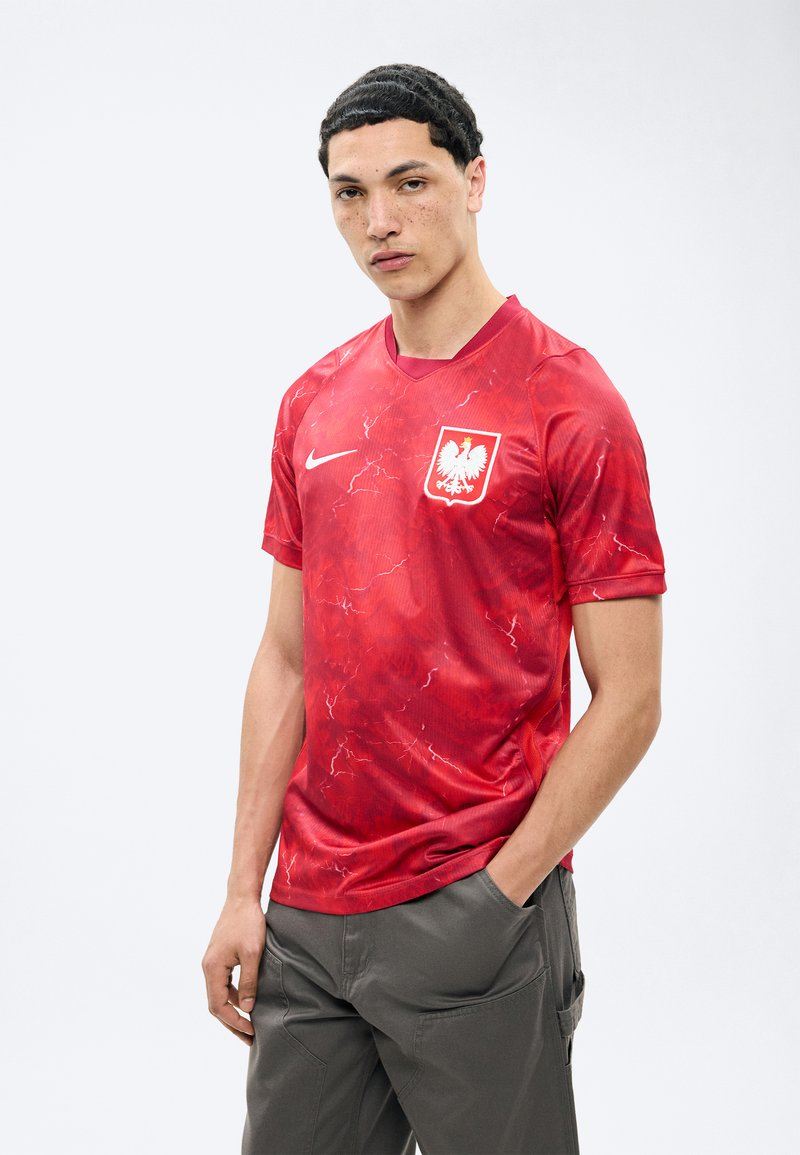 Jeune homme aux cheveux courts et foncés portant un maillot de football rouge de la Pologne avec un blason d'aigle blanc, debout avec une main dans la poche, sur fond blanc.