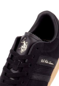Gros plan sur une basket noire avec des lacets noirs, une semelle beige, un logo argenté U.S. Polo Assn. et le nom de la marque brodé sur le côté.