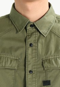 Camicia verde oliva realizzata in misto cotone, con colletto a punta, bottoni a pressione neri e due tasche sul petto in tessuto testurizzato.