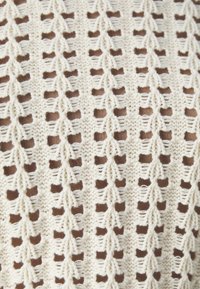 Dzianinowy top w kolorze kremowym, z wzorem ażurowym, posiadający naprzemiennie ułożone grupy małych, trójkątnych kształtów oraz jednolite brązowe akcenty poniżej.