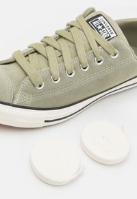 Zapatillas de ante verde con suela de goma negra y blanca, que presentan ojales plateados y cordones a juego, acompañadas de dos cordones redondos blancos.