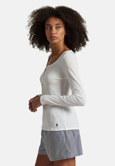 Vrouw met krullend haar, gekleed in een wit, langärmelig, met knopen voorzien top en blauw-wit gestreepte korte broek, staand zijwaarts en naar links kijkend.