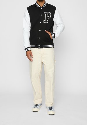 Mand iført sort og hvid varsityjakke med bogstavet P, beige bukser og hvide sneakers, stående med den ene hånd i jakkelommen.