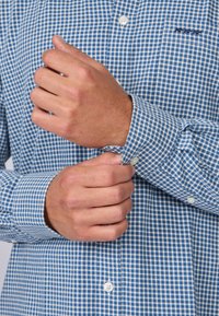 Chemise à carreaux bleue et blanche à boutons avec col, présentant des manches longues et des boutons blancs. Gros plan sur les poignets en train d'être boutonnés.