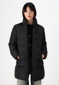 Melnā pūkainā jaka ar augstu apkakli, taisnstūrveida quilted dizainu, sānu kabatām ar rāvējslēdzēju un gludu tekstūru. Uzvilkta virs tumša džempera.