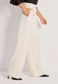 Wijde, hooggetailleerde broek in crème stof met voorplooien en zijzakken. Gecombineerd met zwarte, puntige hakken voor contrast.