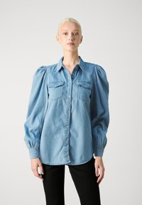 Világoskék farmering puffos ujjal, gombos elejével és két zsebbel a mellrészen. Az ujjak ráncoltak, ami texturált részletet ad az ingnek. Fekete nadrággal viselve.