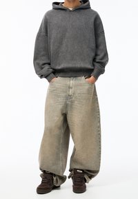 Personne portant un sweat à capuche gris surdimensionné, un jean beige délavé ample et des baskets marron foncé, se tenant avec les mains dans les poches.