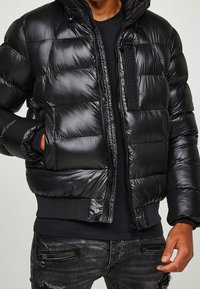 Zwarte pufferjack met glanzende afwerking, ritssluiting aan de voorkant, capuchon en elastische manchetten. Voorzien van steekzakken en merkembleem op de rits.