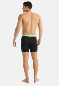 Muž stojící bosý, otočený zády, oblečený v černých boxerkách se zeleným páskem na pozadí čistě bílé.