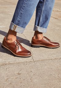 Chaussures habillées en cuir marron avec une finition lisse, des détails cousus et une semelle en caoutchouc. Portées avec un jean en denim bleu clair retroussé.