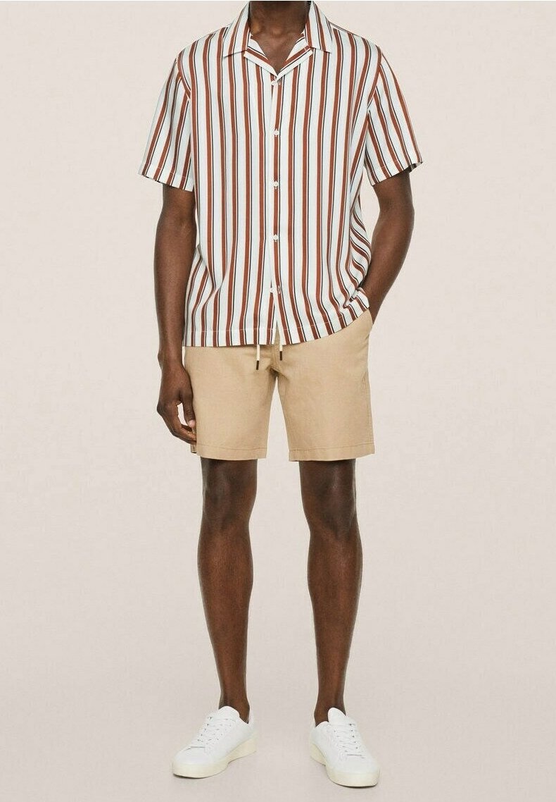 Homme portant une chemise à manches courtes à rayures verticales marron et blanches, un short beige et des baskets blanches, une main dans la poche.