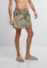 Costumi da bagno da uomo in verde sbiadito con motivo a foglie di palma, vita elastica, tasche laterali, lunghezza sopra il ginocchio, abbinati a infradito neutre.