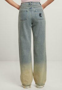 Jeans de mezclilla anchos de color azul claro con un efecto ombré desvanecido. Presenta bolsillos traseros y un parche negro con el logo en el bolsillo derecho. Dobladillo deshilachado.