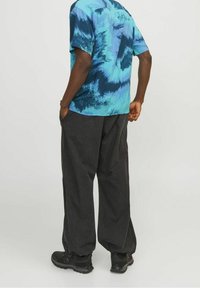 Hombre con zapatos negros, pantalones negros sueltos y una camisa de manga corta azul con estampado, de pie con las manos en los bolsillos y mirando hacia delante.