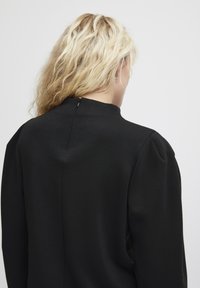Maglione nero con collo alto, maniche a sbuffo e chiusura a chiave sul retro. Il tessuto appare liscio con una leggera trama.