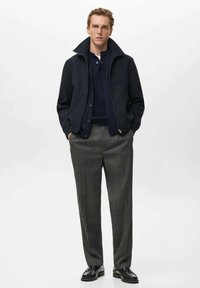 Giacca zip grigia scuro con colletto rialzato sopra un maglione blu navy, abbinata a pantaloni grigi su misura e scarpe nere. Texture piatta.