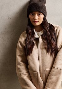Beige Wildlederjacke mit Reißverschluss auf der Vorderseite, die zwei große Taschen und einen kragen aus weißem Fleece hat. Dazu wird eine dunkelbraune Strickmütze getragen.