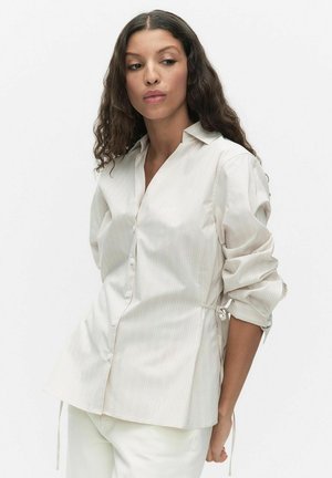 Pitkäkiharainen nainen pukeutuneena valkoiseen, ohuin raitoin koristeltuun nappipaitaan, jonka hihat on kääritty, ja valkoisiin housuihin, seisoo yksinkertaisen taustan edessä.