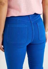 Jeans de denim azul con una textura suave, dos bolsillos traseros y un diseño ajustado. Llevados con una blusa de color rosa claro, que muestra parte de la cinturilla.