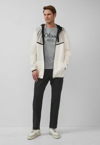 Biała kurtka na zamek błyskawiczny z czarnymi akcentami, szara koszulka z grafiką, czarne spodnie i białe trampki w beżowe paski, stojąca na szarym tle.