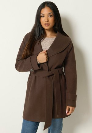 Femme aux longs cheveux foncés portant un manteau marron ceinturé sur un pull beige et un jean bleu, debout devant un fond clair uni.