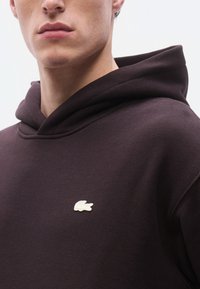 Σκούρα καφέ φούτερ με κουκούλα, που διαθέτει τσέπη καγκουρό και κεντημένο λευκό λογότυπο στο στήθος. Μαλακή υφή με άνετη εφαρμογή και ρυθμιζόμενη κουκούλα.