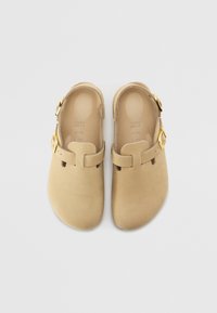 Beige-klomper med mat finish, der har en rund tå, sidebuckle og udskårne ventilationshuller. Glat tekstur og minimalistisk design.