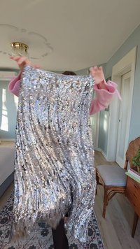 Strieborná látka pokrytá flitrami, s vrstveným strapcovým detailom na spodku. Textúra je lesklá, odráža svetlo v rôznych smeroch.