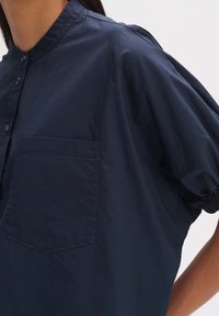 Camisa de manga corta en azul marino de tejido ligero, con cuello alzado, cierre de botones en la parte frontal y un bolsillo de parche en el lado izquierdo.