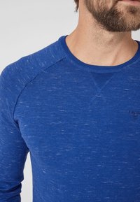 Maglietta a maniche lunghe blu con collo a girocollo e una miscela di texture, caratterizzata da maniche raglan e sottili punte di luce. Piccolo logo sul petto.