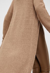 Langer, gerippter, beiger Cardigan mit seitlichen Schlitzen, kombiniert mit braunen, figurschmeichelnden Hosen. Das Material wirkt weich und strukturiert.