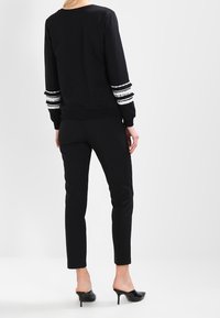 Svart sweatshirt med texturerade vita randiga detaljer på ärmarna, matchad med svarta slim-fit byxor och svarta klackslippers. Slät tyg, avslappnad passform.