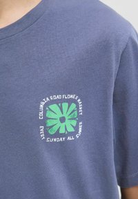 Polo majica v mornarsko modri barvi iz bombaža, ki ima grafiko z zelenim cvetličnim motivom in belim napisom "Cvetlični trg Columbia Road vsako nedeljo poleti."