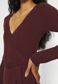 Femme portant une robe pull bordeaux côtelée à manches longues avec un col en V profond et une ceinture nouée à la taille, la main posée sur la hanche.