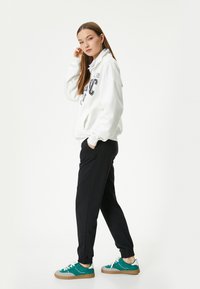 Witte zip-up sweatshirt met grafisch ontwerp, gecombineerd met zwarte tapered joggers en teal sneakers met witte en bruine accenten.