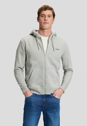 Mężczyzna w jasnoszarej rozpinanej bluzie z kapturem, białym T-shircie i niebieskich dżinsach, stoi z rękami w kieszeniach na jednolitym tle.