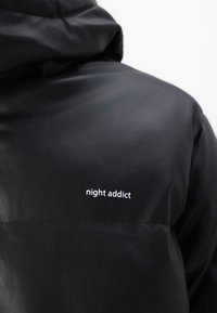 Svart puffjacka med en mjuk yta, huva och texten "night addict" i vitt på baksidan, med en elegant design och inga synliga sömmar.