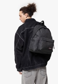 Mochila de tela a rayas en negro y gris con dos bolsillos frontales, correas ajustables y una etiqueta de marca. Llevada en la espalda de una persona.