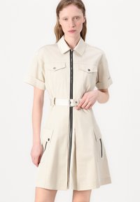 Beige kortärmad klänning i tyg med knytmidja, två bröstfickor och fickor med dragkedja på sidorna, med dragkedja framtill.