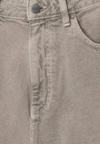 Pantalon en denim beige clair avec une texture lisse, poche avant et coutures contrastantes. Bouton en métal et logo de la marque visibles sur la taille.