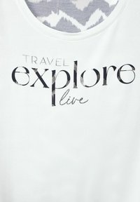 Bílý bavlněný tričko s černým a metalickým potiskem, na kterém je nápis "CESTUJ, objevuj, žij," s kulatým výstřihem a hladkým povrchem.